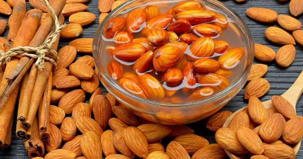 bowl of almonds soaking in water to reduce phytic acid. Almonds are spread on table around it. sticks of cinnamon and a wooden spoon